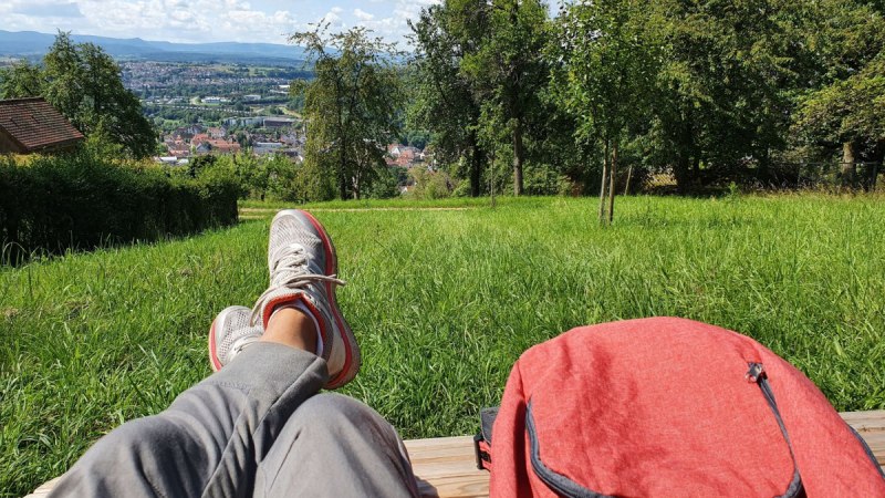 Person entspannt auf einer Bank mit Blick auf eine Stadt in der Ferne, umgeben von grünen Wiesen und Bäumen. Ein roter Rucksack liegt daneben. Person entspannt auf einer Bank mit Blick auf eine Stadt in der Ferne, umgeben von grünen Wiesen und Bäumen. Ein roter Rucksack liegt daneben.