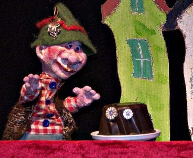 Kasperle-Puppe mit gr&uuml;nem Hut und kariertem Hemd neben einem Schokoladenpudding vor einer gemalten Kulisse mit H&auml;usern., &copy; Bildrechte: Gregor Oehmann