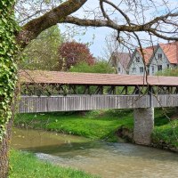Fluss mit überdachter Holzbrücke, am Ufer Büsche und Bäume, auf der rechten Bildseite sind die weißen Giebelseiten und rotbraunen Dachflächen von vier Häusern hinter der Holzbrücke zu erkennen, © Stadt Gaildorf