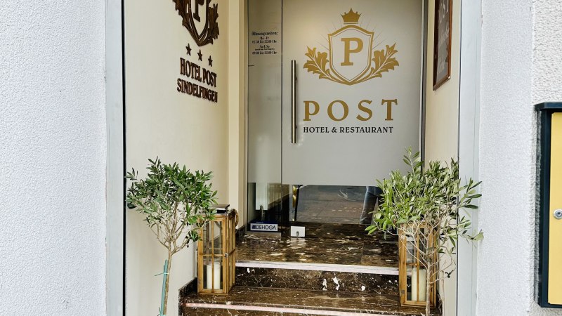 Eingang zum POST Hotel & Restaurant mit Glast&uuml;r, Marmorstufen und Pflanzen in T&ouml;pfen. An der Wand ist das Logo des Hotels zu sehen., &copy; Hotel Post