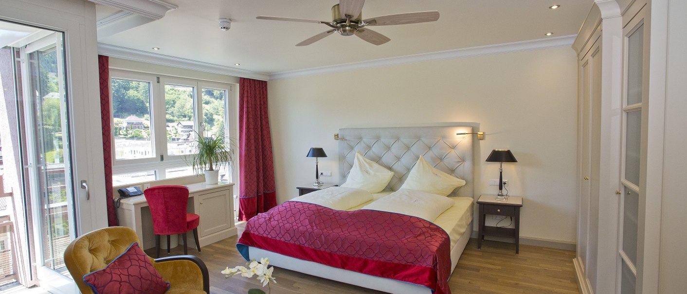 Elegantes Hotelzimmer mit Doppelbett, roten Vorhängen, Balkon und Blick auf grüne Landschaft. Helle Einrichtung mit Deckenventilator und gemütlichen Sitzgelegenheiten., © Mokni's Palais Hotel & Spa Elegantes Hotelzimmer mit Doppelbett, roten Vorhängen, Balkon und Blick auf grüne Landschaft. Helle Einrichtung mit Deckenventilator und gemütlichen Sitzgelegenheiten., © Mokni's Palais Hotel & Spa