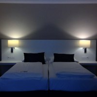 Zwei Einzelbetten mit weißen Laken und schwarzen Kissen in einem Hotelzimmer. An der Wand sind zwei Lampen angebracht, die warmes Licht spenden., © relexa Waldhotel Schatten