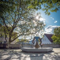 Moderner Brunnen auf gepflastertem Platz, umgeben von Bäumen und Gebäuden. Die Sonne scheint durch die Blätter, der Himmel ist blau mit wenigen Wolken., © René Straube