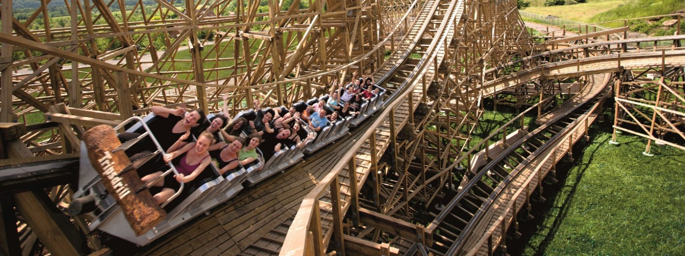 Eine Gruppe von Menschen genie&szlig;t eine Fahrt auf einer Holzachterbahn, w&auml;hrend sie die Arme in die Luft strecken. Die Bahn schl&auml;ngelt sich durch gr&uuml;ne Landschaft., &copy; Erlebnispark Tripsdrill