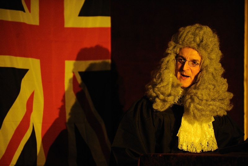Ein Mann in historischer Richterrobe mit Perücke spricht vor einer britischen Flagge. Die Szene ist in warmes Licht getaucht., © Gruseldinner GmbH & Co. KG Ein Mann in historischer Richterrobe mit Perücke spricht vor einer britischen Flagge. Die Szene ist in warmes Licht getaucht., © Gruseldinner GmbH & Co. KG