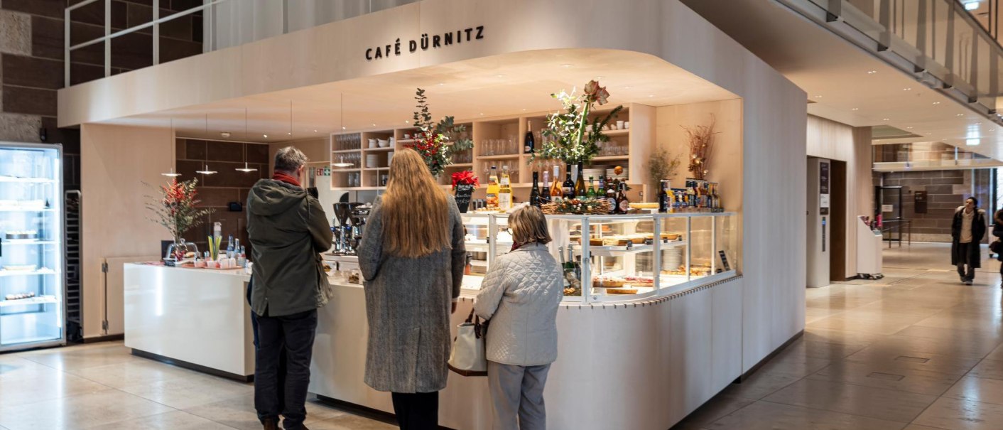 Theke mit Getr&auml;nken und Kuchenvitrine, dekoriert mit Pflanzen. Drei Personen stehen an., &copy; SMG, Sarah Schmid