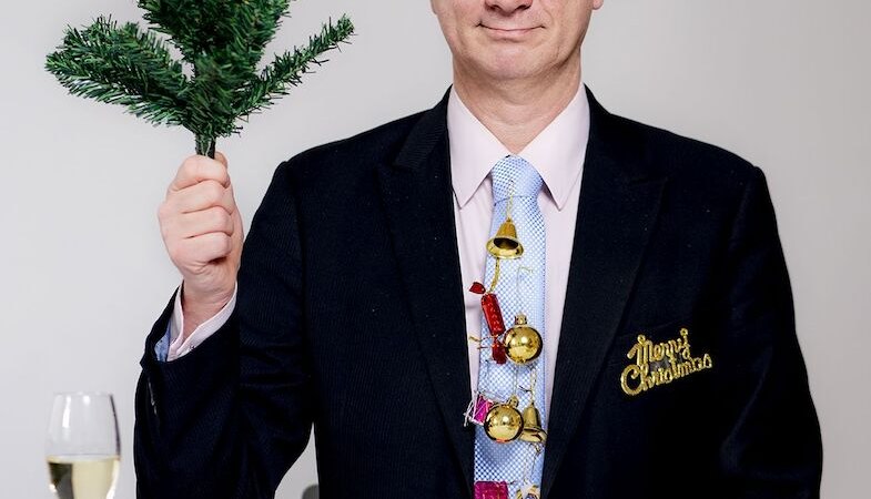 Mann in Anzug mit Weihnachtsdeko auf der Krawatte, hält einen Tannenzweig. Auf dem Tisch stehen ein Sektglas und Snacks., © Stadthalle Leonberg
