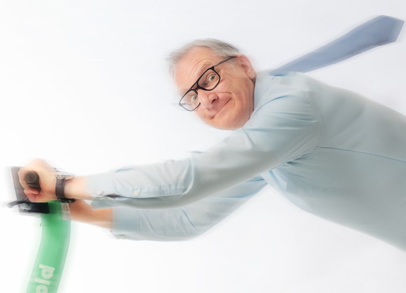 Ein Mann mit Brille und blauer Krawatte hält sich an einem grünen E-Scooter fest. Er ist dynamisch zur Seite geneigt, was Bewegung suggeriert., © Mauerwerk GmbH