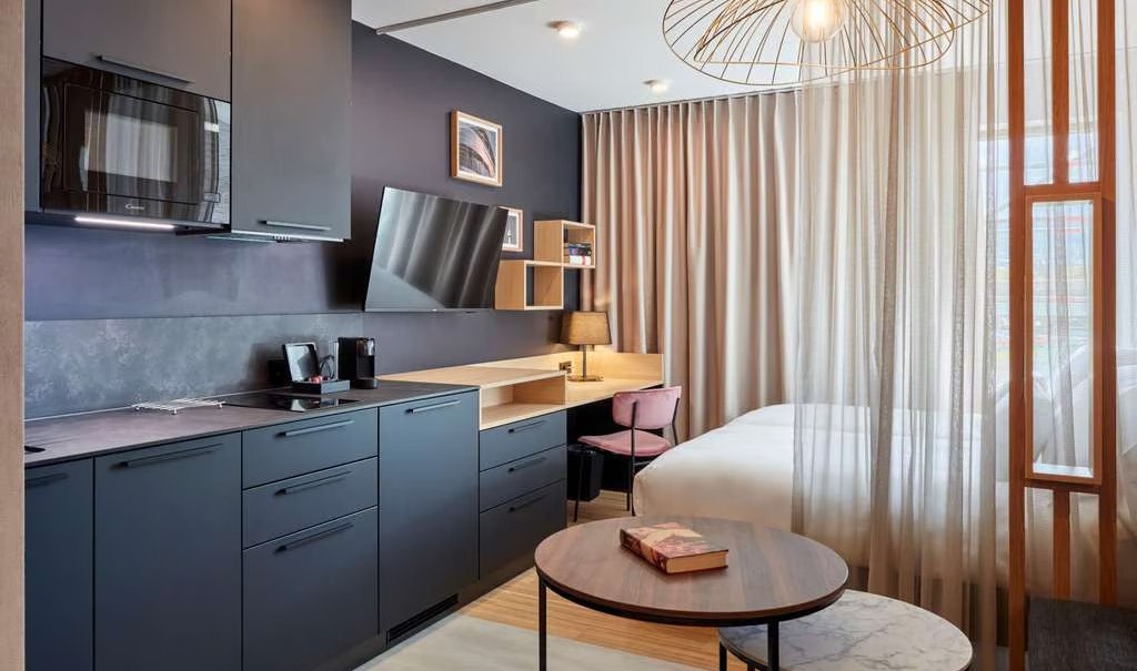 Modernes Studio mit dunkler Küche, Schreibtisch, Bett und stilvoller Beleuchtung. Vorhänge trennen den Schlafbereich ab., © Adagio Modernes Studio mit dunkler Küche, Schreibtisch, Bett und stilvoller Beleuchtung. Vorhänge trennen den Schlafbereich ab., © Adagio