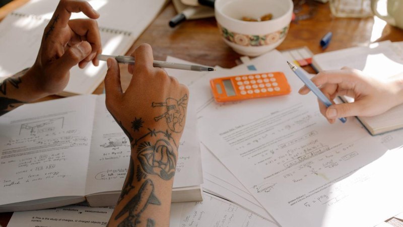 Zwei Personen bearbeiten mathematische Aufgaben mit Taschenrechner, Notizen und B&uuml;chern auf einem Tisch. Eine Person hat t&auml;towierte Arme., &copy; MGBH Pliensauvorstadt