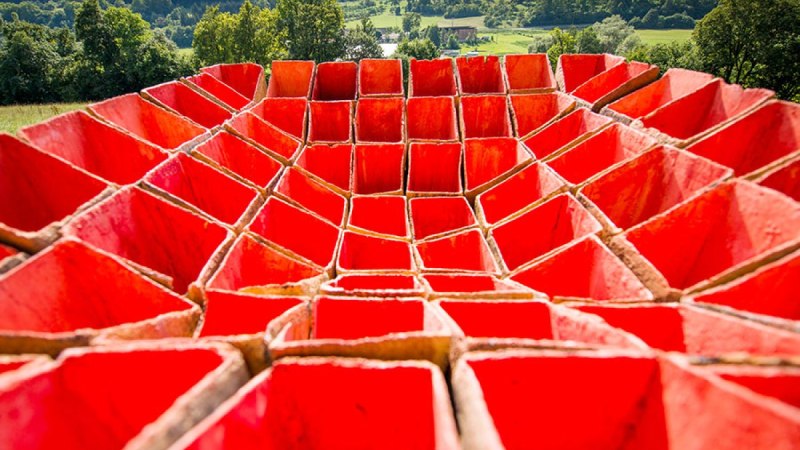 Rote, geometrische Struktur in Form eines Kraters vor einer gr&uuml;nen Landschaft mit B&auml;umen und H&uuml;geln im Hintergrund., &copy; Natur.Nah. Sch&ouml;nbuch & Heckeng&auml;u