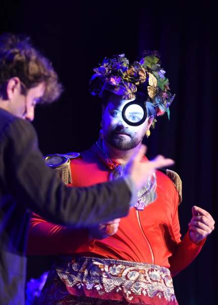 Ein Mann in rotem Kostüm mit Blumenkrone und Monokel steht auf einer Bühne, während ein anderer Mann im Vordergrund gestikuliert., © Sascha von Donat Ein Mann in rotem Kostüm mit Blumenkrone und Monokel steht auf einer Bühne, während ein anderer Mann im Vordergrund gestikuliert., © Sascha von Donat