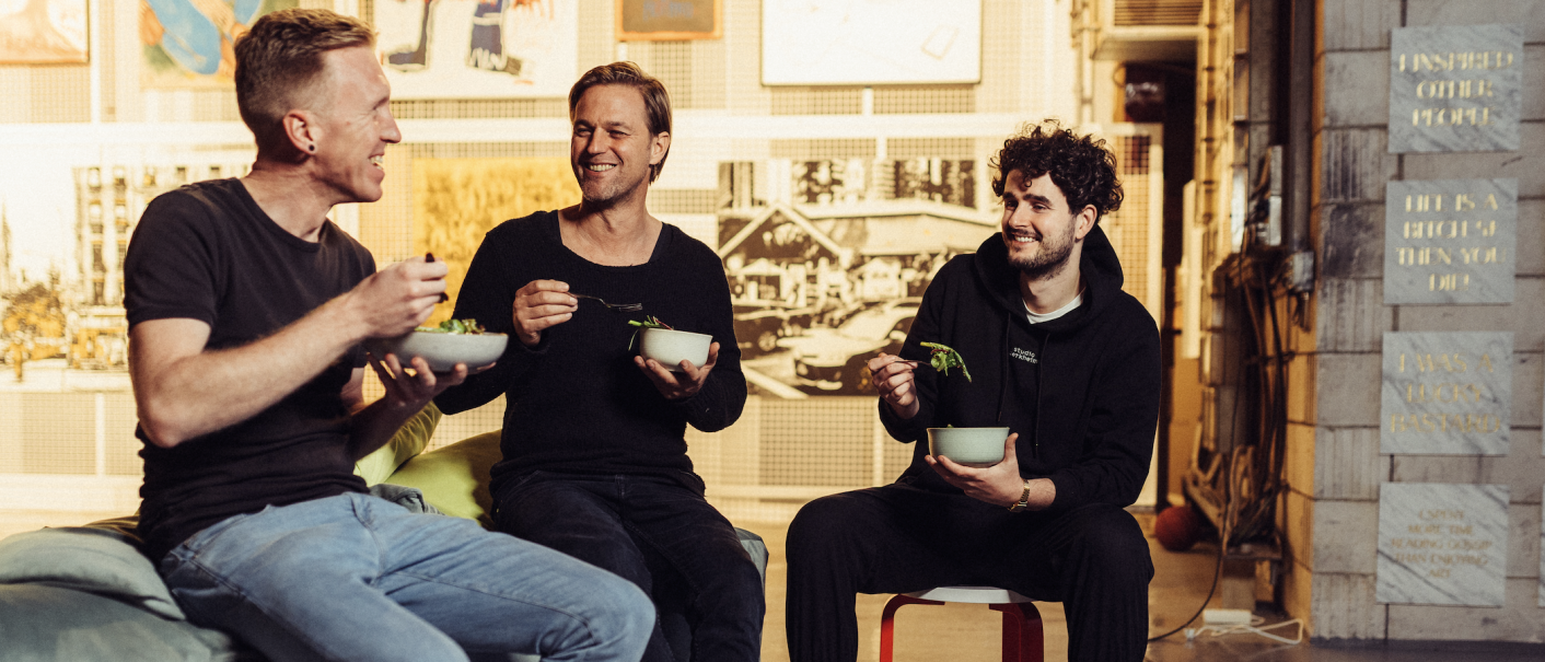 Drei Männer sitzen lachend zusammen und essen Salat in einem gemütlichen, künstlerisch gestalteten Raum., © Vhy! GmbH Drei Männer sitzen lachend zusammen und essen Salat in einem gemütlichen, künstlerisch gestalteten Raum., © Vhy! GmbH