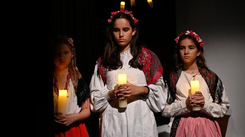 Drei M&auml;dchen in traditioneller Kleidung mit Blumenkr&auml;nzen halten Kerzen in einem dunklen Raum. Sie wirken konzentriert und ernst., &copy; Kunstschule Labyrinth