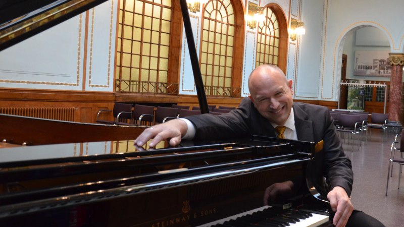 Ein Mann in einem Anzug lehnt sich l&auml;chelnd an einen Steinway-Fl&uuml;gel in einem eleganten Raum mit hohen Decken und verzierten Fenstern., &copy; Tim Kandel