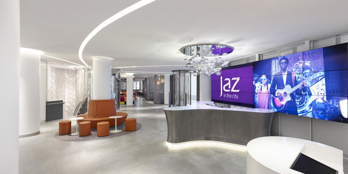 Moderne Rezeption mit geschwungenem Tresen, großem Bildschirm mit "Jaz in the City"-Logo und stilvoller Beleuchtung., © Jaz Hotel Stuttgart