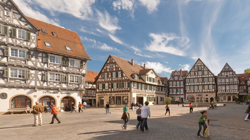 Der Marktplatz in Schorndorf zeigt malerische Fachwerkhäuser unter blauem Himmel. Menschen spazieren und verweilen auf dem Platz., © Oswald Der Marktplatz in Schorndorf zeigt malerische Fachwerkhäuser unter blauem Himmel. Menschen spazieren und verweilen auf dem Platz., © Oswald