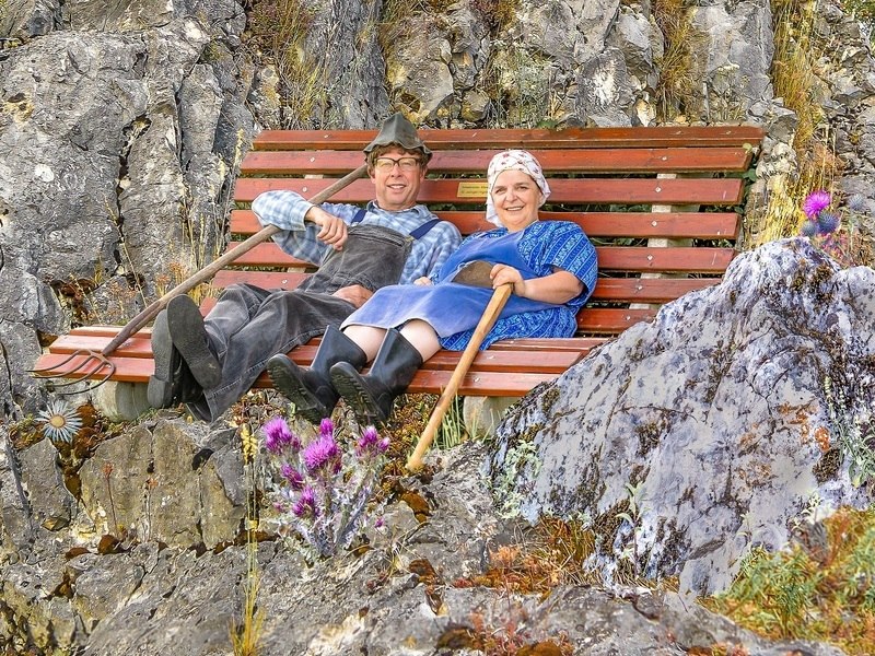 Zwei Personen in traditioneller Kleidung sitzen entspannt auf einer Bank vor einer felsigen Landschaft. Sie halten Wanderstöcke und lächeln in die Kamera., © NPG Messe und Veranstaltungs-GmbH Zwei Personen in traditioneller Kleidung sitzen entspannt auf einer Bank vor einer felsigen Landschaft. Sie halten Wanderstöcke und lächeln in die Kamera., © NPG Messe und Veranstaltungs-GmbH