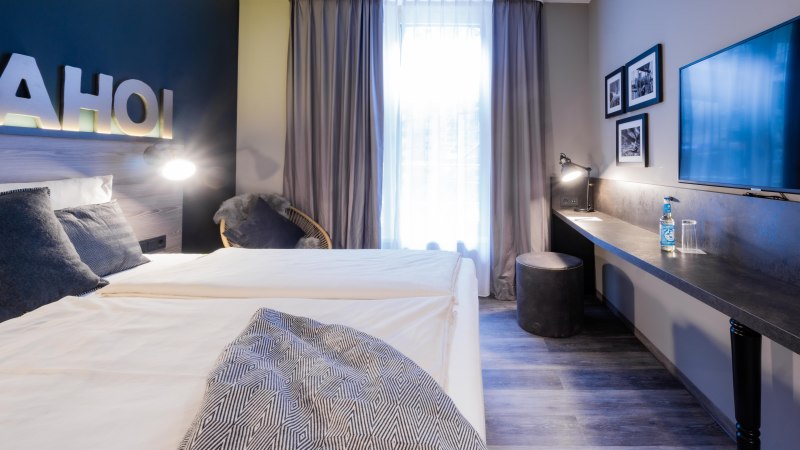 Modernes Zimmer mit Doppelbett, Schreibtisch, Flachbildfernseher und Dekoration. Schriftzug 'AHOI' an der Wand, neutrale Farben und gemütliche Atmosphäre., © HARBR. hotel Ludwigsburg / DQuadrat Living GmbH