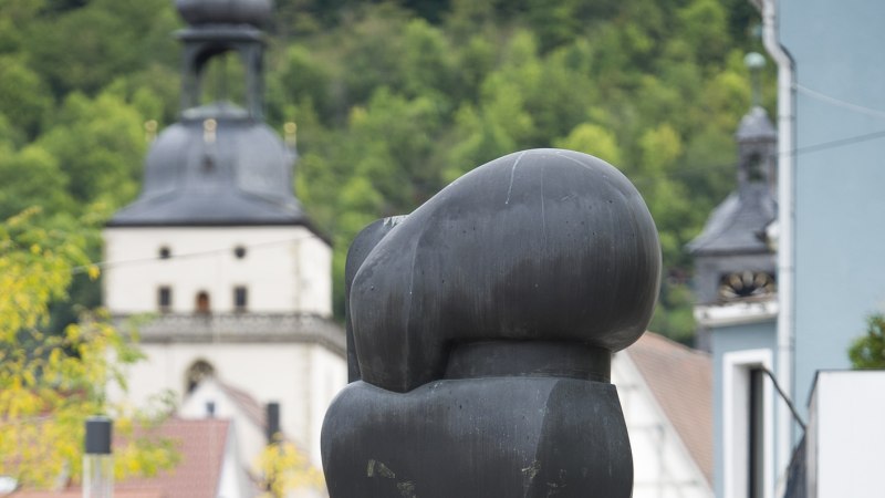 Moderne Skulptur in einer st&auml;dtischen Umgebung, im Hintergrund ein Kirchturm und gr&uuml;ne B&auml;ume. Die Skulptur ist abstrakt und dunkel gef&auml;rbt., &copy; Archiv W&uuml;rth