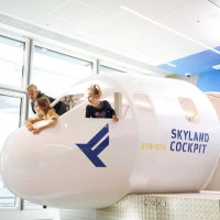 Drei Kinder spielen in einem Cockpit-Nachbau mit der Aufschrift 'SkyLand Cockpit' und blicken aus den Fenstern. Helle, moderne Umgebung., © Flughafen Stuttgart