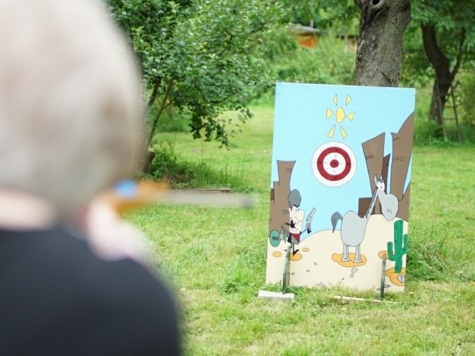 Person zielt mit Blasrohr auf eine bemalte Holztafel mit Zielscheibe im Freien., © Cool-Tours StattReisen Person zielt mit Blasrohr auf eine bemalte Holztafel mit Zielscheibe im Freien., © Cool-Tours StattReisen