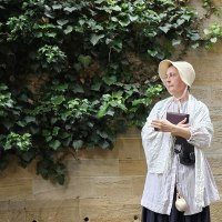 Eine Frau in historischer Kleidung steht nachdenklich vor einer mit Efeu bewachsenen Steinmauer. Sie h&auml;lt ein Buch in der Hand., &copy; Xenia Busam