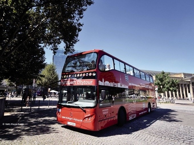 Roter Doppeldeckerbus der Stuttgart Citytour auf einem gepflasterten Platz, umgeben von Bäumen und Gebäuden., © SMG, Pierre Polak Roter Doppeldeckerbus der Stuttgart Citytour auf einem gepflasterten Platz, umgeben von Bäumen und Gebäuden., © SMG, Pierre Polak