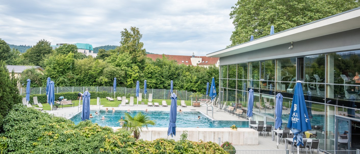 Ein Reha-Zentrum mit Pool, Liegestühlen und blauen Sonnenschirmen. Im Hintergrund sind Bäume und Gebäude zu sehen., © Christophsbad Klinikgruppe Ein Reha-Zentrum mit Pool, Liegestühlen und blauen Sonnenschirmen. Im Hintergrund sind Bäume und Gebäude zu sehen., © Christophsbad Klinikgruppe