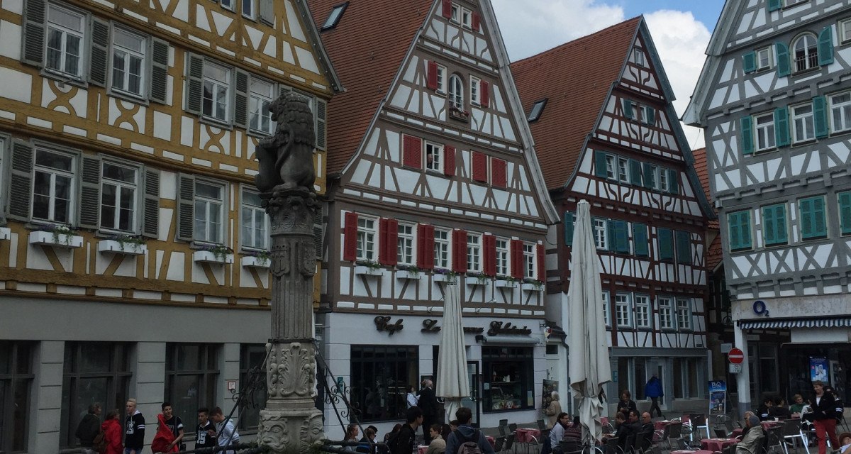 Fachwerkhäuser am Marktplatz in Herrenberg, Deutschland. Ein Brunnen steht im Vordergrund, umgeben von Menschen und Cafétischen. Der Himmel ist blau mit Wolken., © www.pro-cycl.de