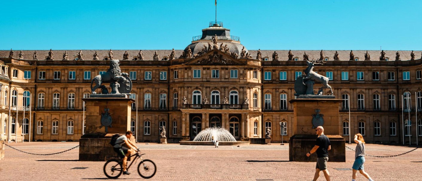 Das Neue Schloss Stuttgart bei sonnigem Wetter. Menschen gehen und ein Fahrradfahrer fährt im Vordergrund. Zwei Statuen flankieren den Eingang., © Stuttgart-Marketing GmbH, Sarah Schmid Das Neue Schloss Stuttgart bei sonnigem Wetter. Menschen gehen und ein Fahrradfahrer fährt im Vordergrund. Zwei Statuen flankieren den Eingang., © Stuttgart-Marketing GmbH, Sarah Schmid