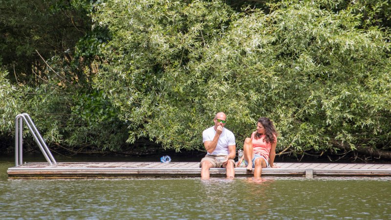 Zwei Personen sitzen auf einem Steg am Wasser, umgeben von üppigem Grün. Sie genießen die Natur im Naherholungsgebiet Bürgerseen Kirchheim unter Teck., © Stadt Kirchheim unter Teck Zwei Personen sitzen auf einem Steg am Wasser, umgeben von üppigem Grün. Sie genießen die Natur im Naherholungsgebiet Bürgerseen Kirchheim unter Teck., © Stadt Kirchheim unter Teck
