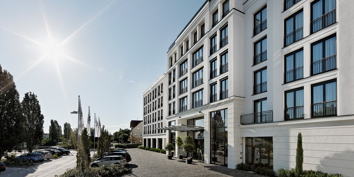 Moderne Gebäudefassade mit großen Fenstern, sonniger Himmel, Autos und Flaggen im Vordergrund., © Parkhotel Stuttgart Messe-Airport