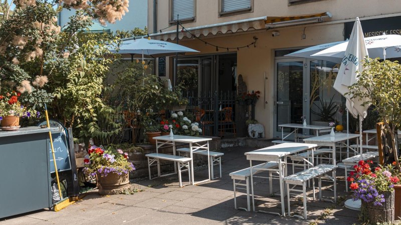 Gemütlicher Außenbereich eines Cafés mit weißen Tischen und Stühlen, umgeben von üppigen Pflanzen und bunten Blumen., © Stuttgart Marketing GmbH, Sarah Schmid Gemütlicher Außenbereich eines Cafés mit weißen Tischen und Stühlen, umgeben von üppigen Pflanzen und bunten Blumen., © Stuttgart Marketing GmbH, Sarah Schmid