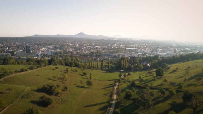 Gr&uuml;ne Felder erstrecken sich vor einer Stadt, die von H&uuml;geln umgeben ist. Die Szenerie wirkt ruhig und idyllisch.