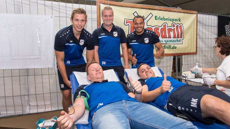 Zwei Männer spenden Blut, während drei weitere in Sportkleidung daneben stehen und lächeln. Eine Frau betreut die Blutspende., © Erlebnispark Tripsdrill Zwei Männer spenden Blut, während drei weitere in Sportkleidung daneben stehen und lächeln. Eine Frau betreut die Blutspende., © Erlebnispark Tripsdrill