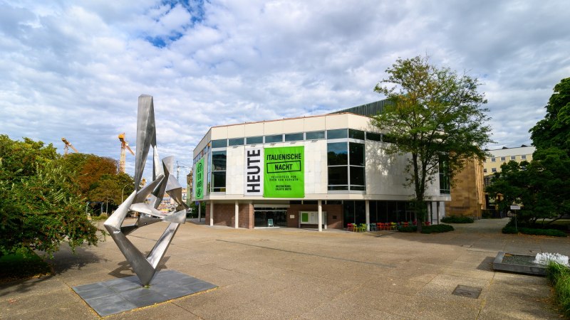 Moderne Skulptur vor dem Schauspielhaus mit großen Fenstern und Plakaten für 'Italienische Nacht'. Wolkenbedeckter Himmel, Bäume und Baukräne im Hintergrund., © Björn Klein Moderne Skulptur vor dem Schauspielhaus mit großen Fenstern und Plakaten für 'Italienische Nacht'. Wolkenbedeckter Himmel, Bäume und Baukräne im Hintergrund., © Björn Klein