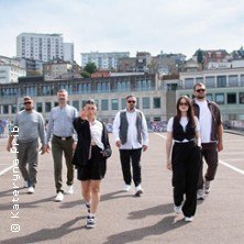 Sechs Personen gehen in l&auml;ssiger Kleidung auf einer Stra&szlig;e. Im Hintergrund sind Geb&auml;ude und ein blauer Himmel zu sehen., &copy; links im Bild
