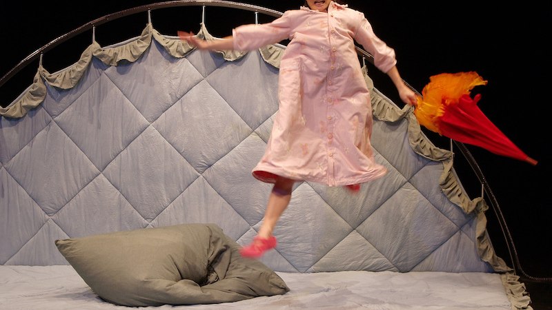 Eine Person in einem rosa Schlafanzug springt auf einem gro&szlig;en Bett. Sie h&auml;lt einen roten Regenschirm und tr&auml;gt eine blaue M&uuml;tze., &copy; FILharmonie Filderstadt