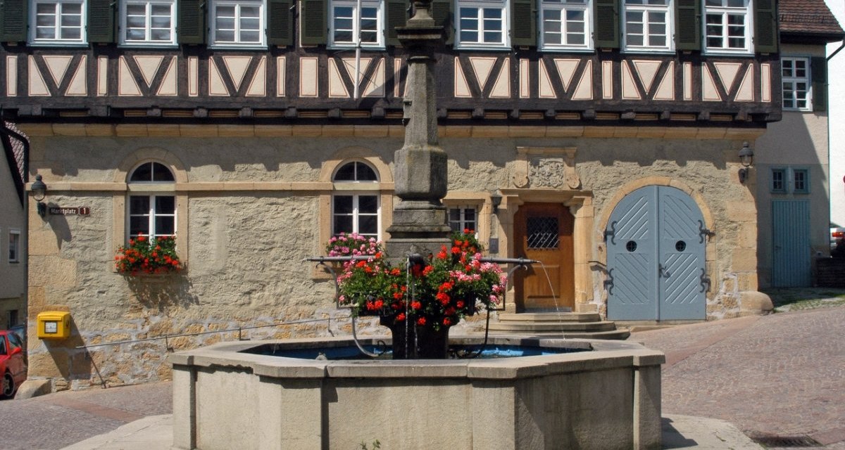 Fachwerkhaus mit Brunnen und Blumen im Vordergrund, gelber Briefkasten an der Wand, T&uuml;r mit Verzierungen, Fensterl&auml;den ge&ouml;ffnet., &copy; Natur.Nah. Sch&ouml;nbuch & Heckeng&auml;u