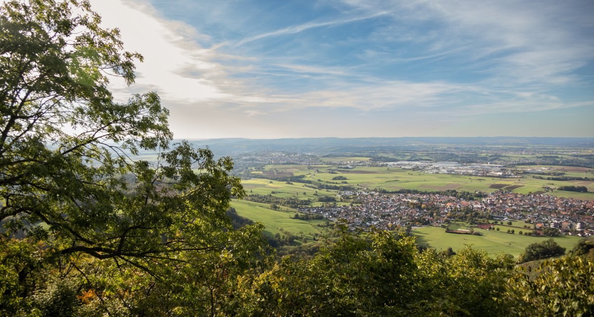 Blick vom Aussichtspunkt Hoher Fels auf eine weite Landschaft mit Feldern, einem Dorf und bewölktem Himmel. Bäume rahmen das Bild ein., © Foto Thomas Zehnder Blick vom Aussichtspunkt Hoher Fels auf eine weite Landschaft mit Feldern, einem Dorf und bewölktem Himmel. Bäume rahmen das Bild ein., © Foto Thomas Zehnder