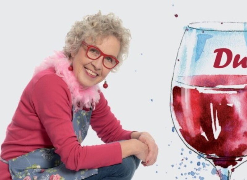 Eine Frau mit lockigem Haar, roter Brille und rosa Schal posiert lächelnd neben einer großen Weinglas-Illustration., © Kulturfenster Bönnigheim e. V. Eine Frau mit lockigem Haar, roter Brille und rosa Schal posiert lächelnd neben einer großen Weinglas-Illustration., © Kulturfenster Bönnigheim e. V.