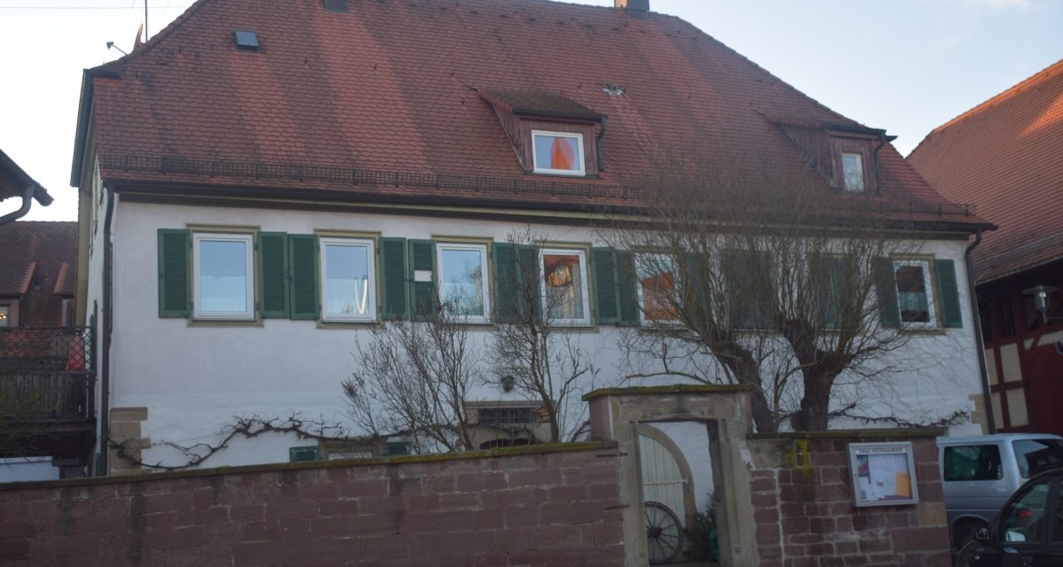 Ein zweistöckiges Gebäude mit rotem Ziegeldach und grünen Fensterläden hinter einer Steinmauer. Ein Torbogen führt in den Innenhof., © Land der 1000 Hügel - Kraichgau-Stromberg Ein zweistöckiges Gebäude mit rotem Ziegeldach und grünen Fensterläden hinter einer Steinmauer. Ein Torbogen führt in den Innenhof., © Land der 1000 Hügel - Kraichgau-Stromberg