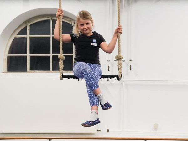 Ein Kind sitzt auf einer Schaukel in einem Raum mit Turnmatten und einer großen Fensteröffnung im Hintergrund., © Kunstschule Labyrinth Ein Kind sitzt auf einer Schaukel in einem Raum mit Turnmatten und einer großen Fensteröffnung im Hintergrund., © Kunstschule Labyrinth