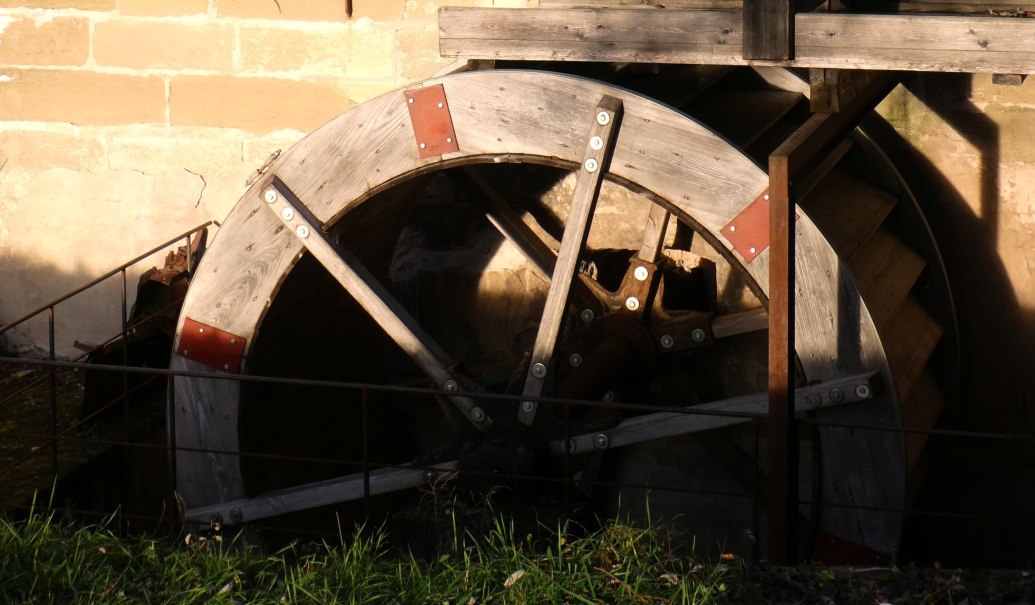 Ein altes h&ouml;lzernes M&uuml;hlrad mit Metallverst&auml;rkungen, das an einer Steinmauer befestigt ist. Sonnenlicht f&auml;llt auf das Rad., &copy; Stadtmarketing Leonberg