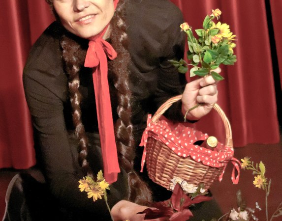 Eine Person im Rotkäppchen-Kostüm hält einen Korb und Blumen vor einem roten Vorhang., © Galli Theater Backnang
