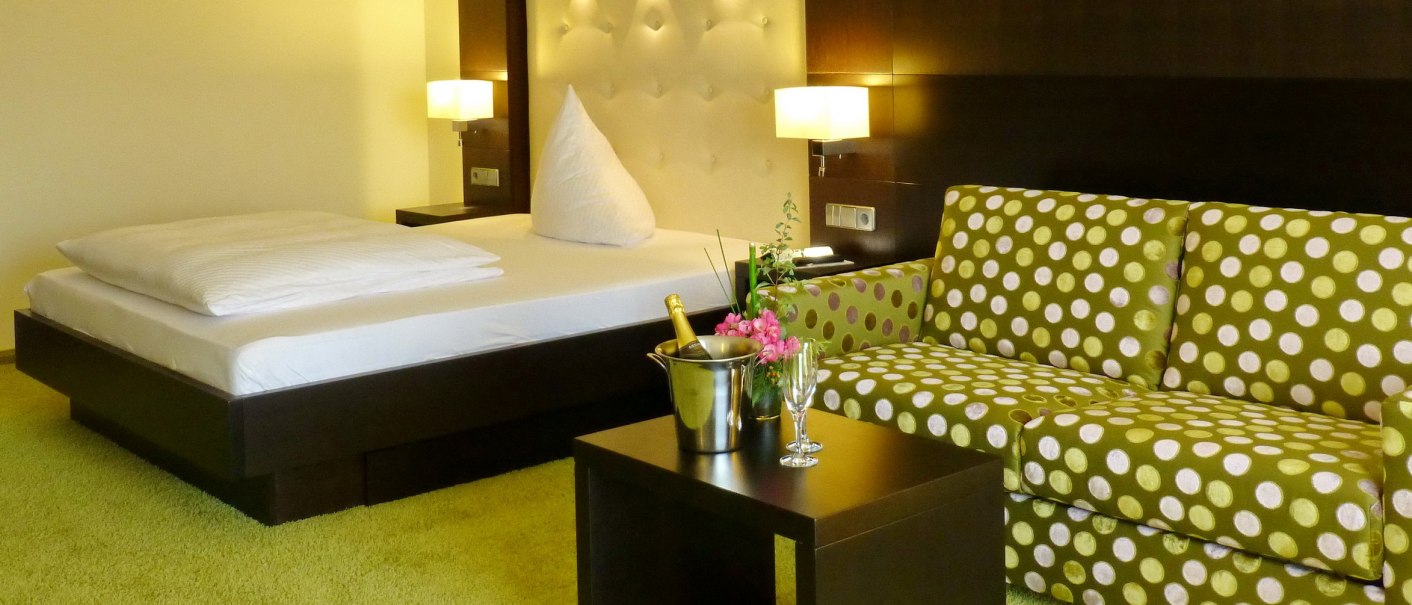 Modernes Hotelzimmer mit Doppelbett, grün gepunktetem Sofa, Tisch mit Sektkühler und Gläsern, auf grünem Teppichboden., © Hotel & Restaurant Sonne