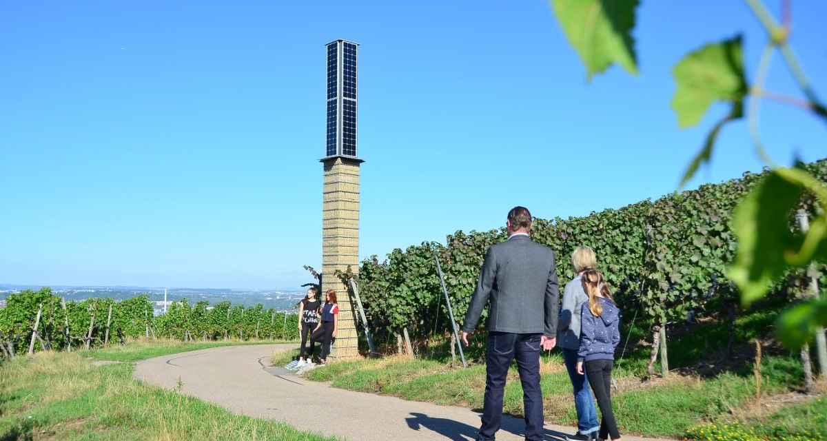 Menschen spazieren auf einem Weg durch die Weinberge in Fellbach. Ein hoher, schmaler Turm steht am Wegesrand. Der Himmel ist klar und blau., &copy; Foto: Simone Mathias