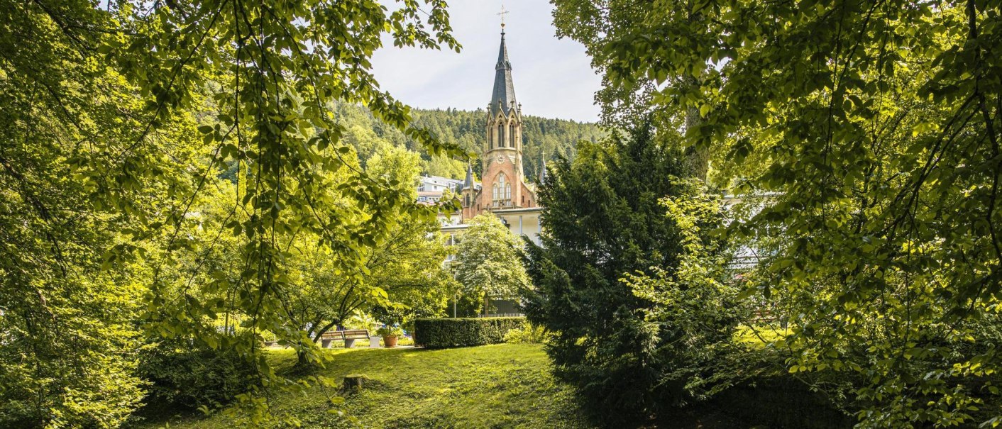 Ein Kirchturm ragt zwischen dichten, gr&uuml;nen B&auml;umen im Kurpark von Bad Wildbad hervor. Die Szene wirkt friedlich und idyllisch., &copy; Stuttgart-Marketing GmbH, Sarah Schmid