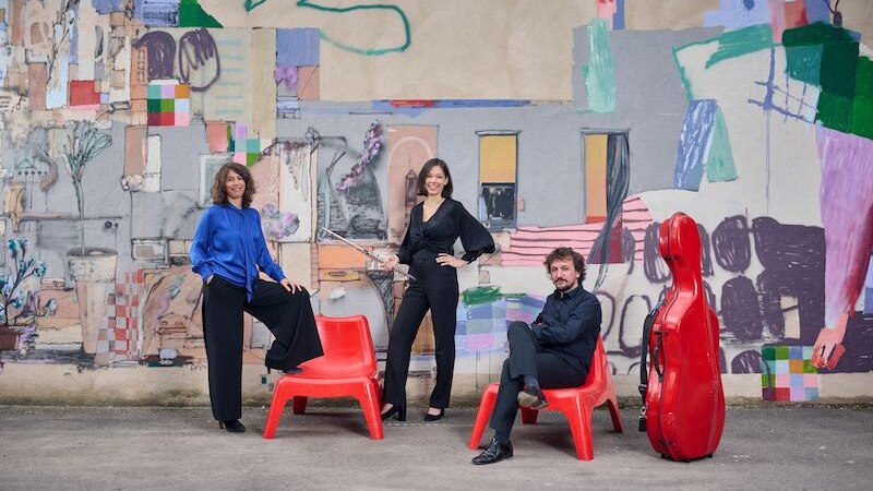 Drei Personen posieren vor einer bunten Wandmalerei. Zwei sitzen auf roten Stühlen, eine steht. Ein roter Cello-Koffer steht daneben., © Stadt Leonberg - Amt für Kultur und Sport